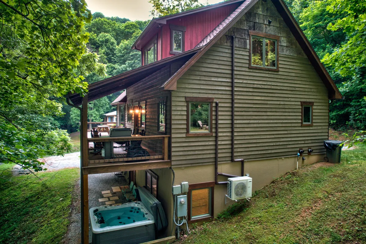 A wide, partially covered wooden deck stretches out amidst a backdrop of lush trees, with a bubbling hot tub tucked to one side. Adirondack and rocking chairs are arranged for relaxation, and the deck offers views of the mountains at sunset. The setting exudes tranquility and connection to nature, perfect for unwinding or entertaining.