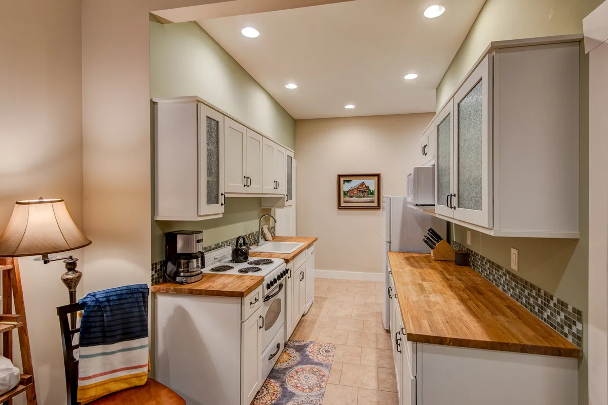 A warm kitchen setting with visible countertops, a stove, refrigerator, wall art, and a glowing lampshade. The arrangement suggests ample space for cooking, socializing, and prepping meals for gatherings or backyard cookouts.