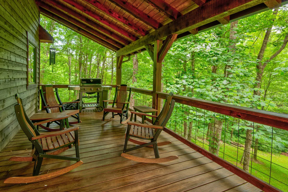A section of the deck features classic rocking chairs and a barbecue grill, with lush greenery providing an intimate, private feel. The space is set up for both relaxation and outdoor cooking, offering views ideal for morning coffee or sunset grilling.