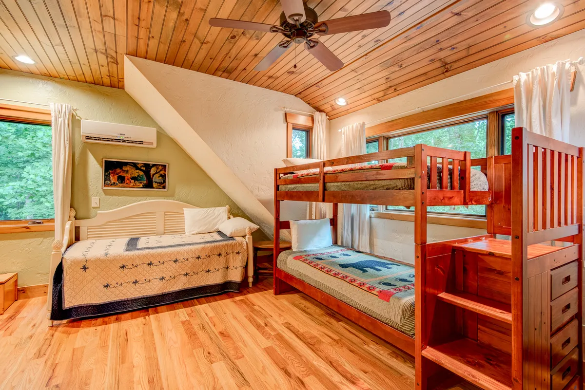 A cozy bunk room features wooden flooring, large windows letting in sunlight, and a cheerful decorative quilt. The space is inviting for kids’ sleepovers, games, and shared laughter, embodying the spirit of family togetherness during relaxed days at home.