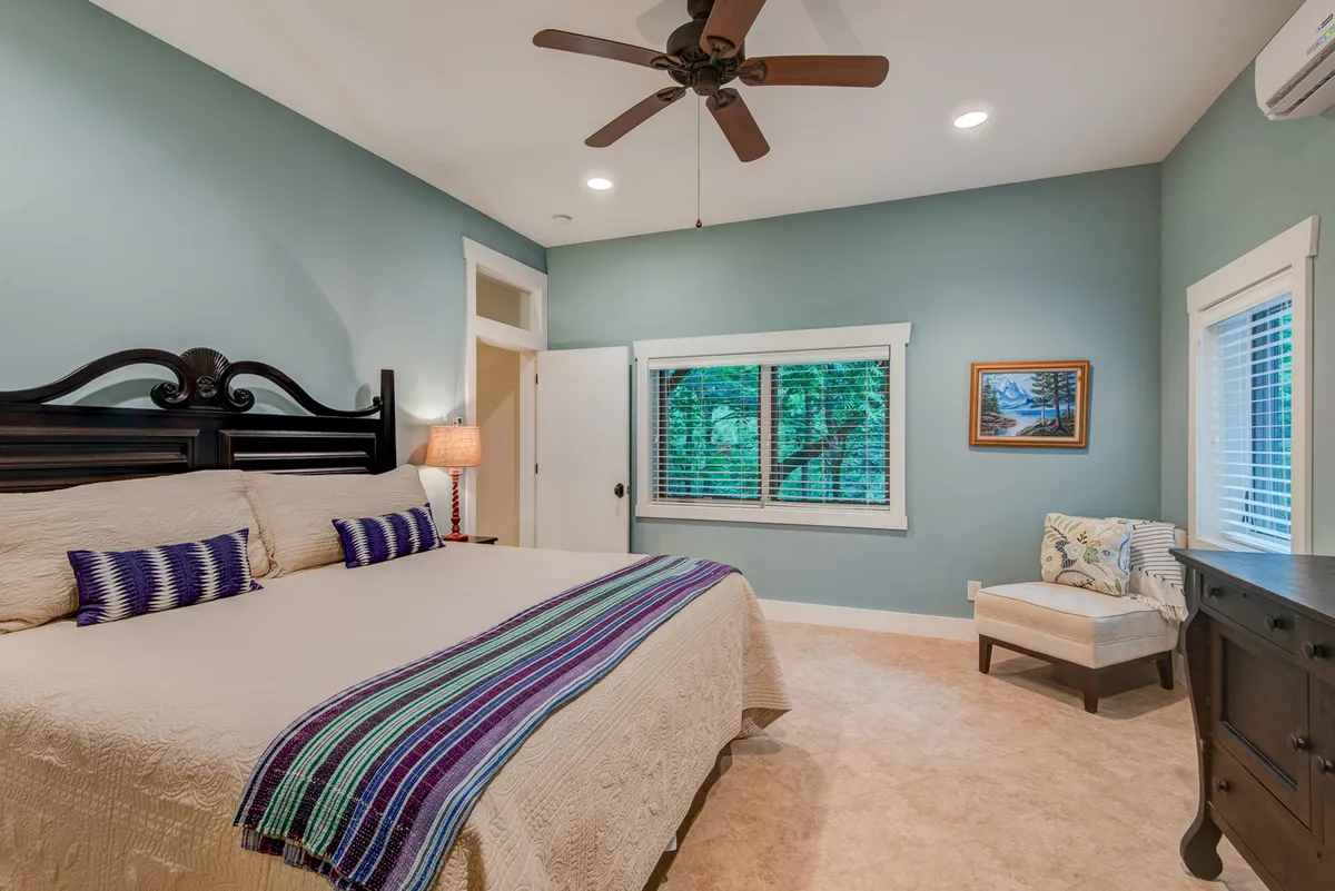 A serene, light-filled bedroom with a king-sized bed dressed in decorative pillows, a comfortable armchair by the window, and framed artwork on the wall. Natural light streams in, and the large windows offer a calming view of greenery outside, highlighting the room's tranquil and private atmosphere.