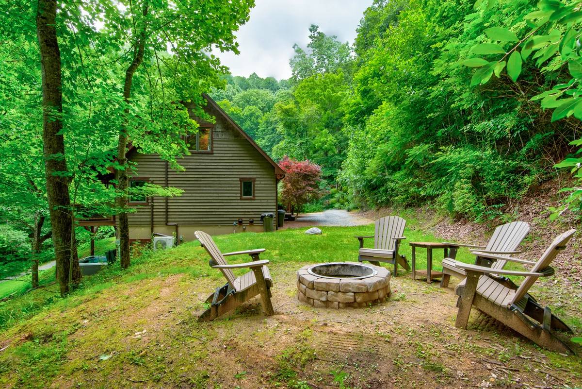 A cozy outdoor fire pit encircled by Adirondack chairs sits against a wooded backdrop. The fire glows as dusk approaches, inviting gatherings for warmth, stories, and marshmallow roasting. The scene radiates camaraderie and comfort in the heart of nature.