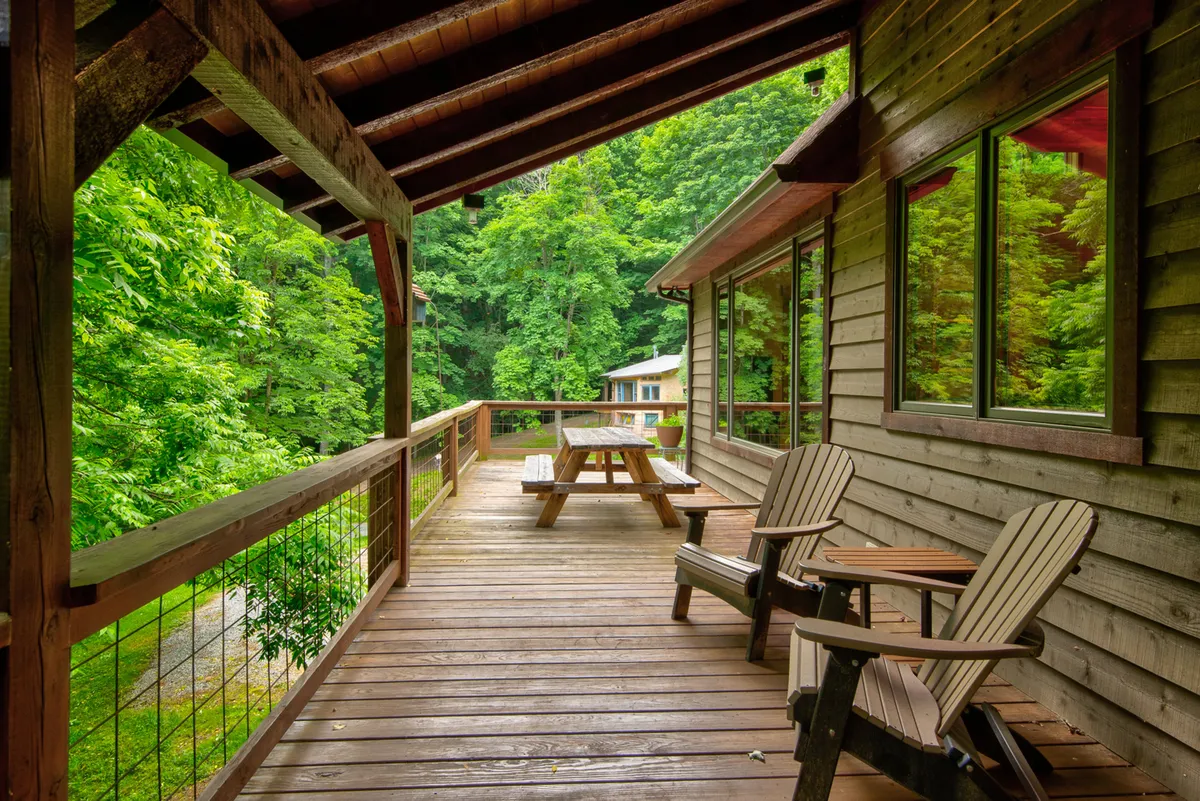 A spacious wooden deck with a picnic table and Adirondack chairs is surrounded by lush greenery. Large windows of the home blur the line between interior and exterior living, offering a seamless connection to the outdoors. The deck is positioned for enjoying both sunrise coffee and evening gatherings under the stars.