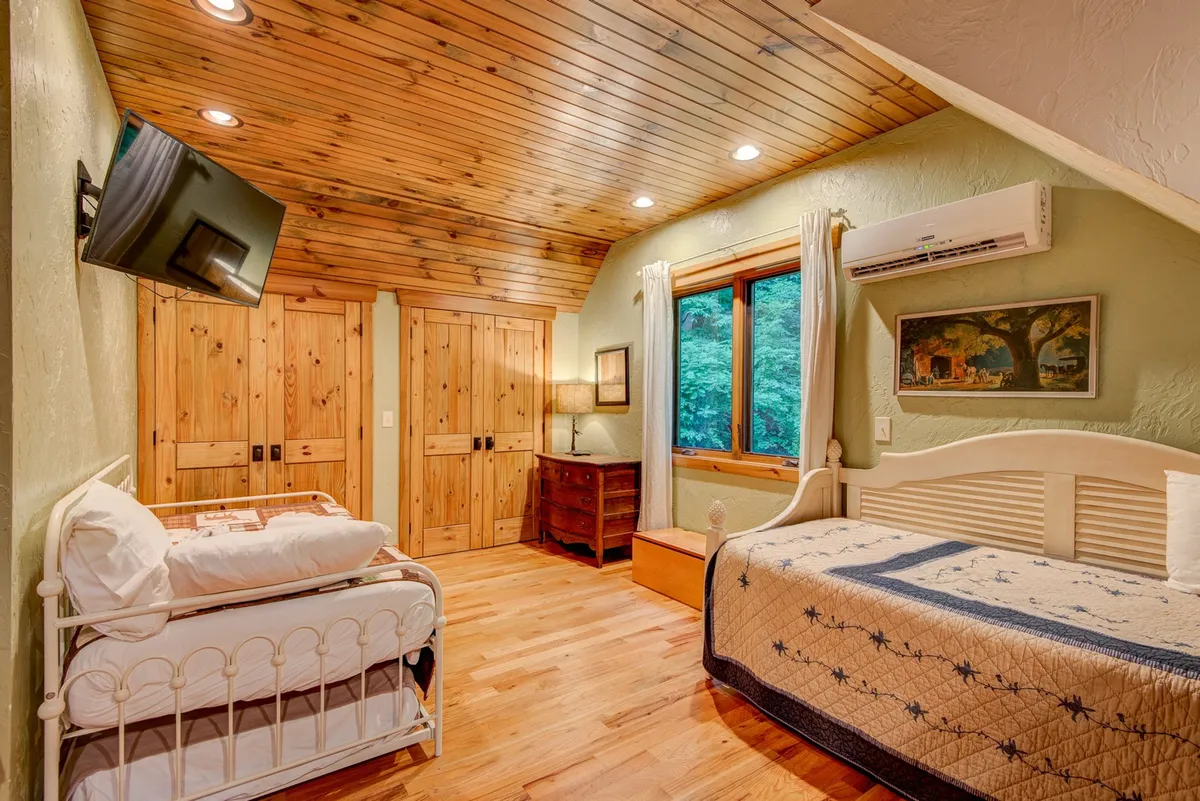 A cozy bedroom with two beds, a rustic wooden ceiling, large window framing outside greenery, and warm wooden furniture. The space is inviting and bright, ideal for either couples or adults seeking privacy, with subtle touches that suggest comfort and thoughtful design.