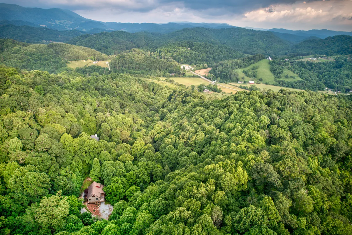 A wide view of the house nestled among expansive green foliage with distant mountains in the background. The setting evokes a sense of peaceful seclusion, ideal for hiking, nature walks, and stargazing. The landscape hints at adventuring opportunities right outside the door, with the home as a cozy refuge after a day of exploration.