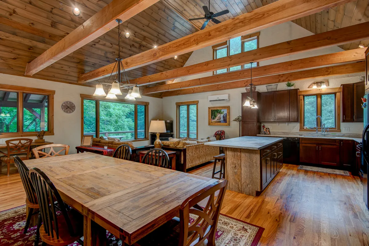 A spacious open-concept interior featuring a large dining table, generous kitchen island, and cozy sofa area. The room is warmed by natural light streaming through large windows, and the rustic wooden beams add character and warmth. The setup invites gatherings, casual dining, and effortless entertaining.