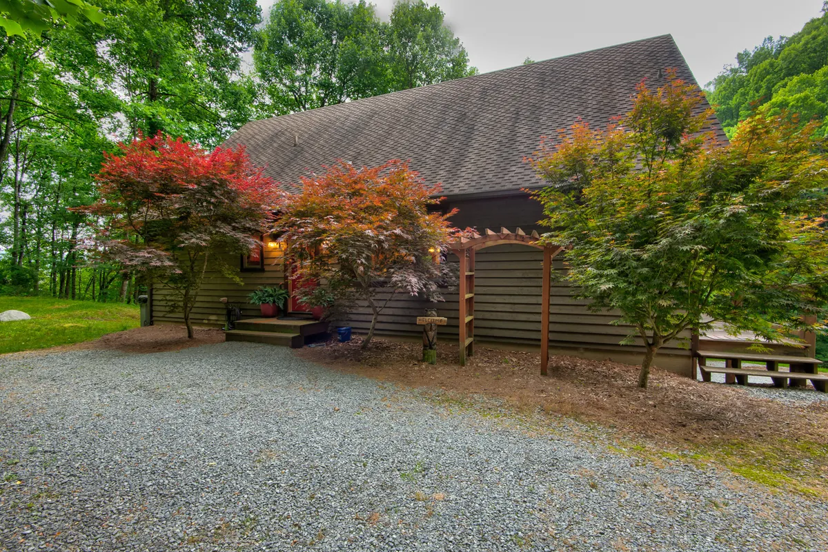 A tranquil exterior shot of the house set against a backdrop of colorful trees, a wide gravel path, and a spacious porch area. The scene suggests plenty of open land surrounding the home, perfect for outdoor activities and relaxation.