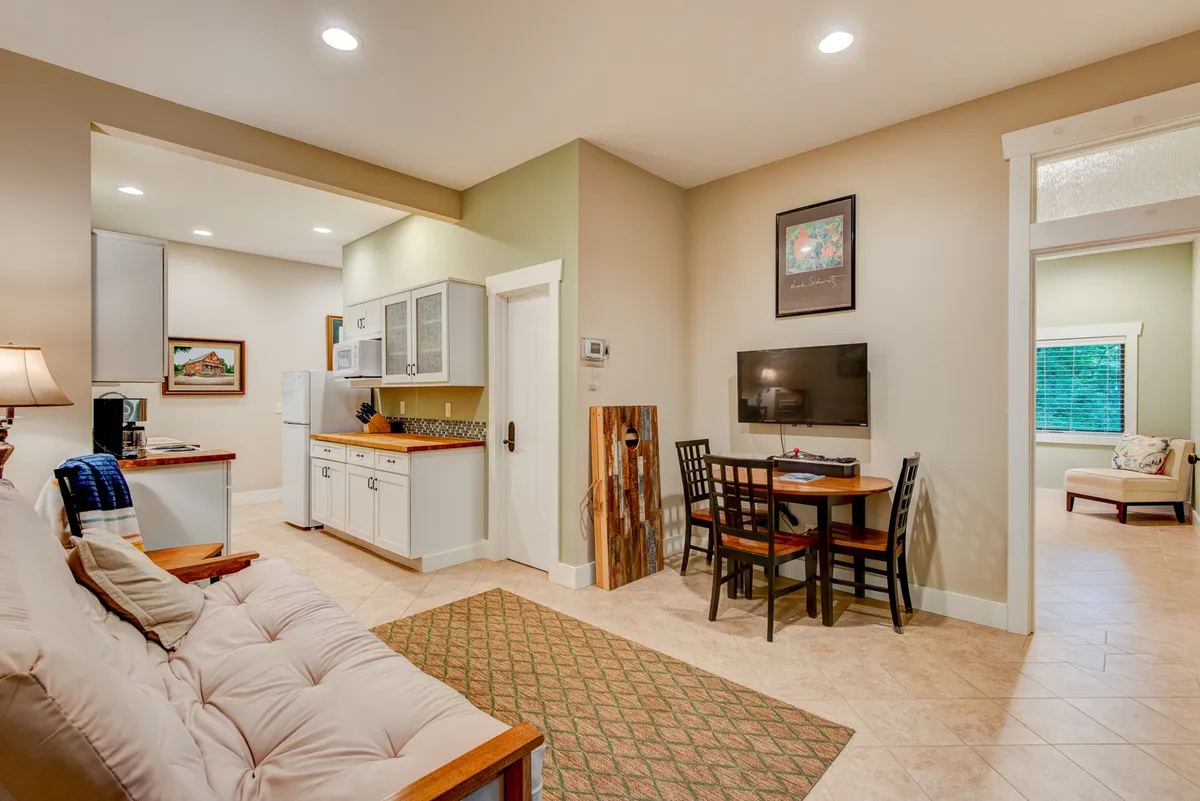 The image displays a welcoming open-plan space featuring a sofa, sleek dining table, and a kitchen area with visible cabinetry and wall art. The kitchen is immediately adjacent to the dining area, suggesting a communal, flexible environment ideal for group meals and casual entertainment.