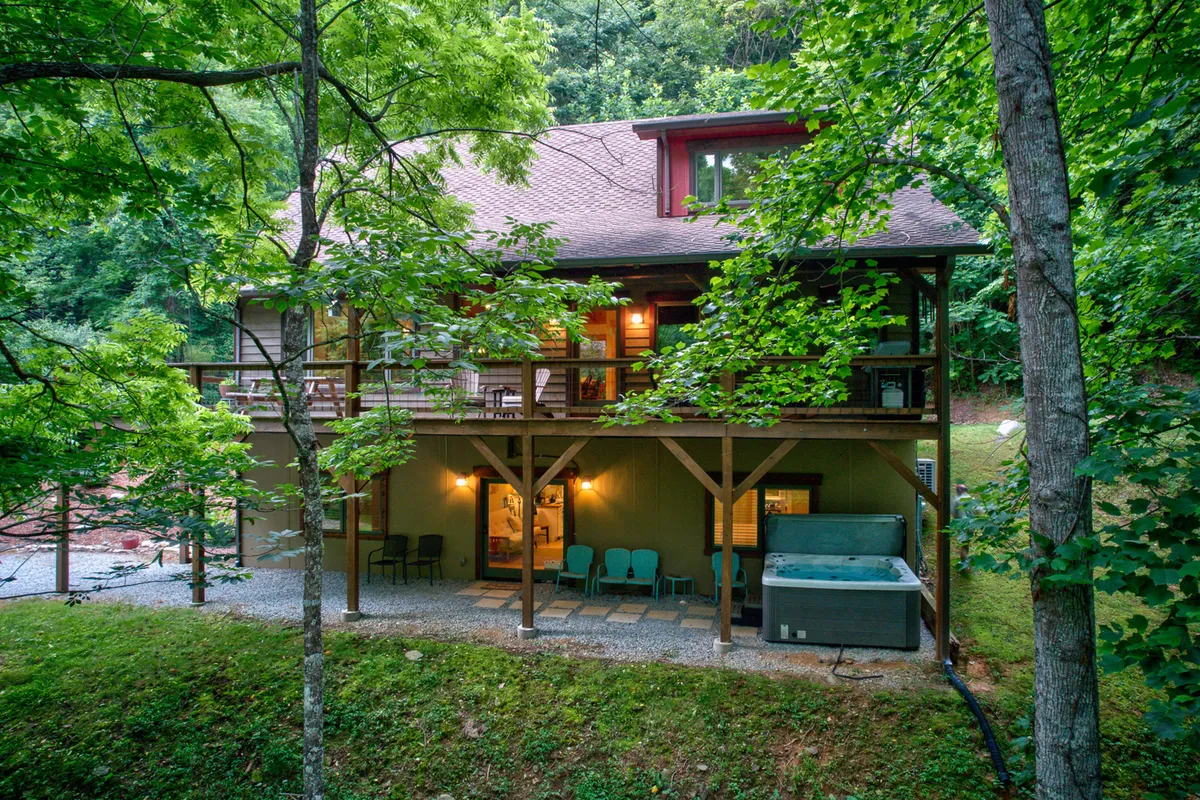 A spacious wooden deck features a relaxing hot tub and ample outdoor seating, all surrounded by lush trees. The setting is tranquil, with plenty of room for groups to gather or individuals to unwind privately. The area feels inviting for both daytime and evening activities, such as soaking in the hot tub under the stars or sharing drinks with friends at sunset.