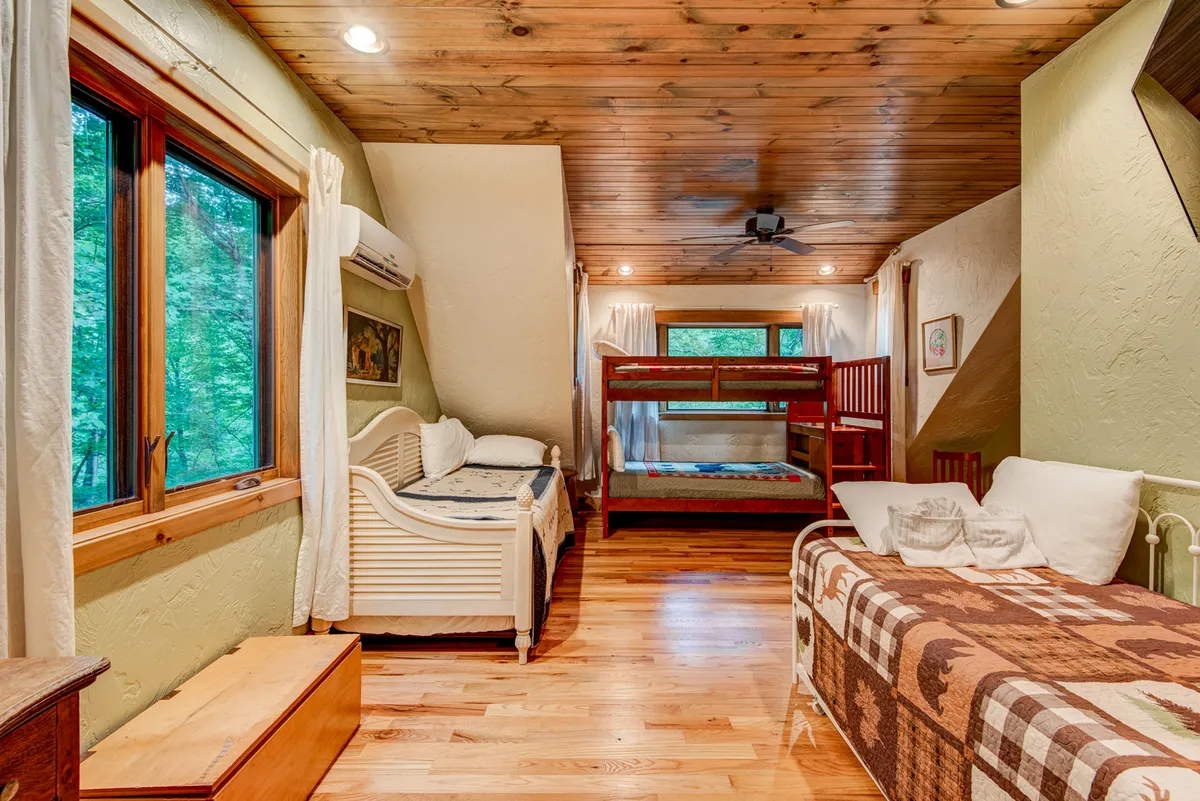 A vibrant upstairs bunk room with multiple bunk beds, a daybed, wood-paneled ceiling, and large windows filling the space with sunlight. The room is playful yet spacious, designed for children or cousins to sleep, play games, and bond, capturing the essence of a kid haven.