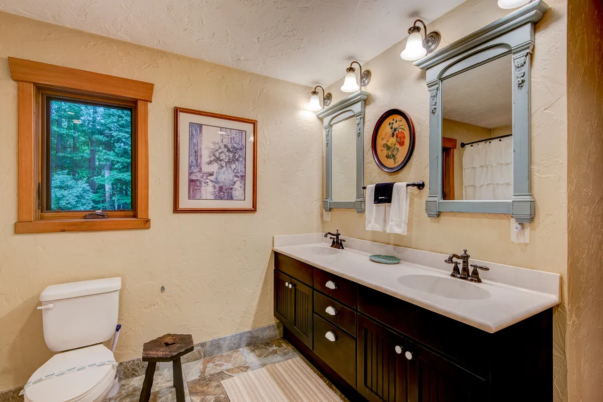 A bright, calming ensuite bathroom featuring a double vanity with mirrors, a large window overlooking greenery, and elegant wall art. The space is designed for relaxation and personal care, with fresh morning light enhancing its spa-like ambiance.