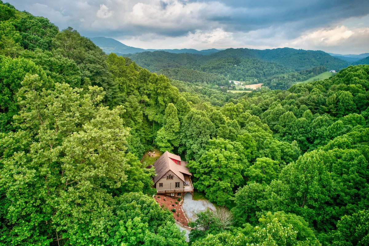 This serene exterior image showcases The Guf nestled amid lush trees with a dramatic mountain backdrop, capturing the natural beauty and tranquil setting that make it an ideal retreat for nature lovers and hikers. The porch offers an inviting spot to enjoy the fresh air and panoramic views.